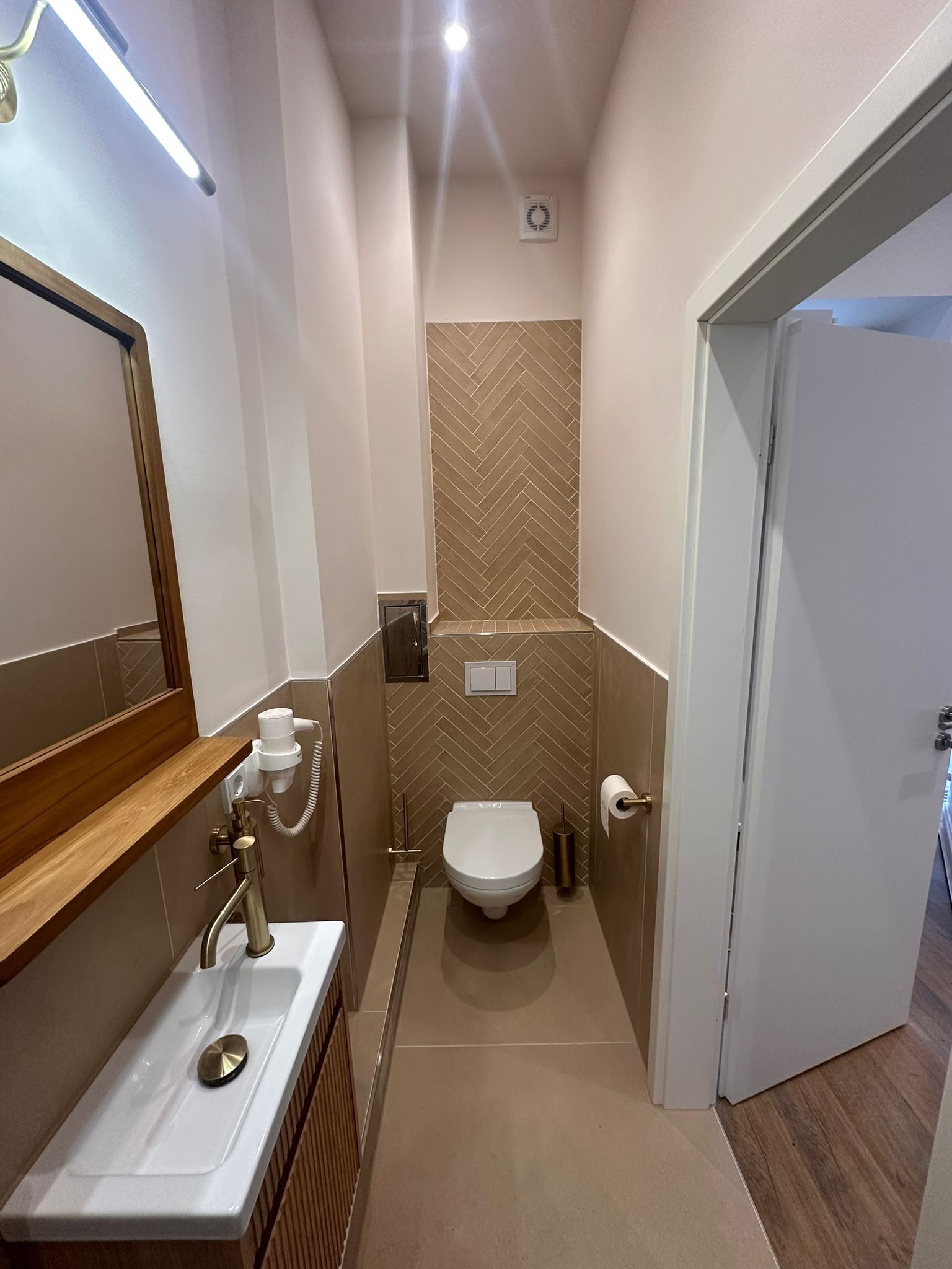 Fertiges schmales Badezimmer: Wand-WC vor einer Fläche aus beigen Fischgrätfliesen, links ein Waschtisch mit Holzplatte und Spiegel, Armaturen in Messingfarbe, fugenloser Bodenbelag.