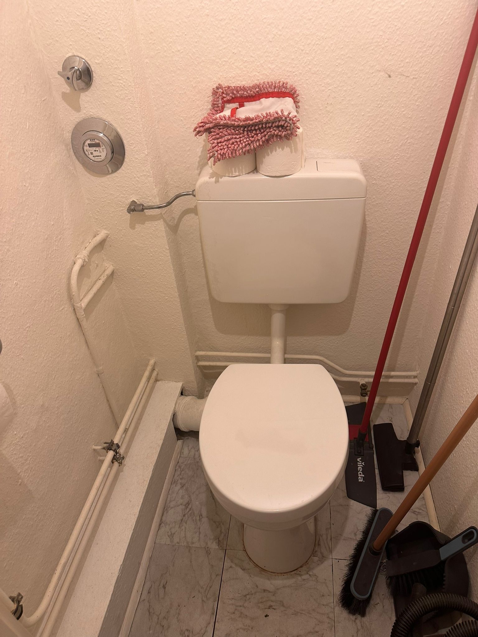Enges WC vor der Sanierung: freistehende Keramik mit Aufputzspülkasten, Leitungen sichtbar an der Wand, Marmorfliesen am Boden, Putzgerät in der Ecke.