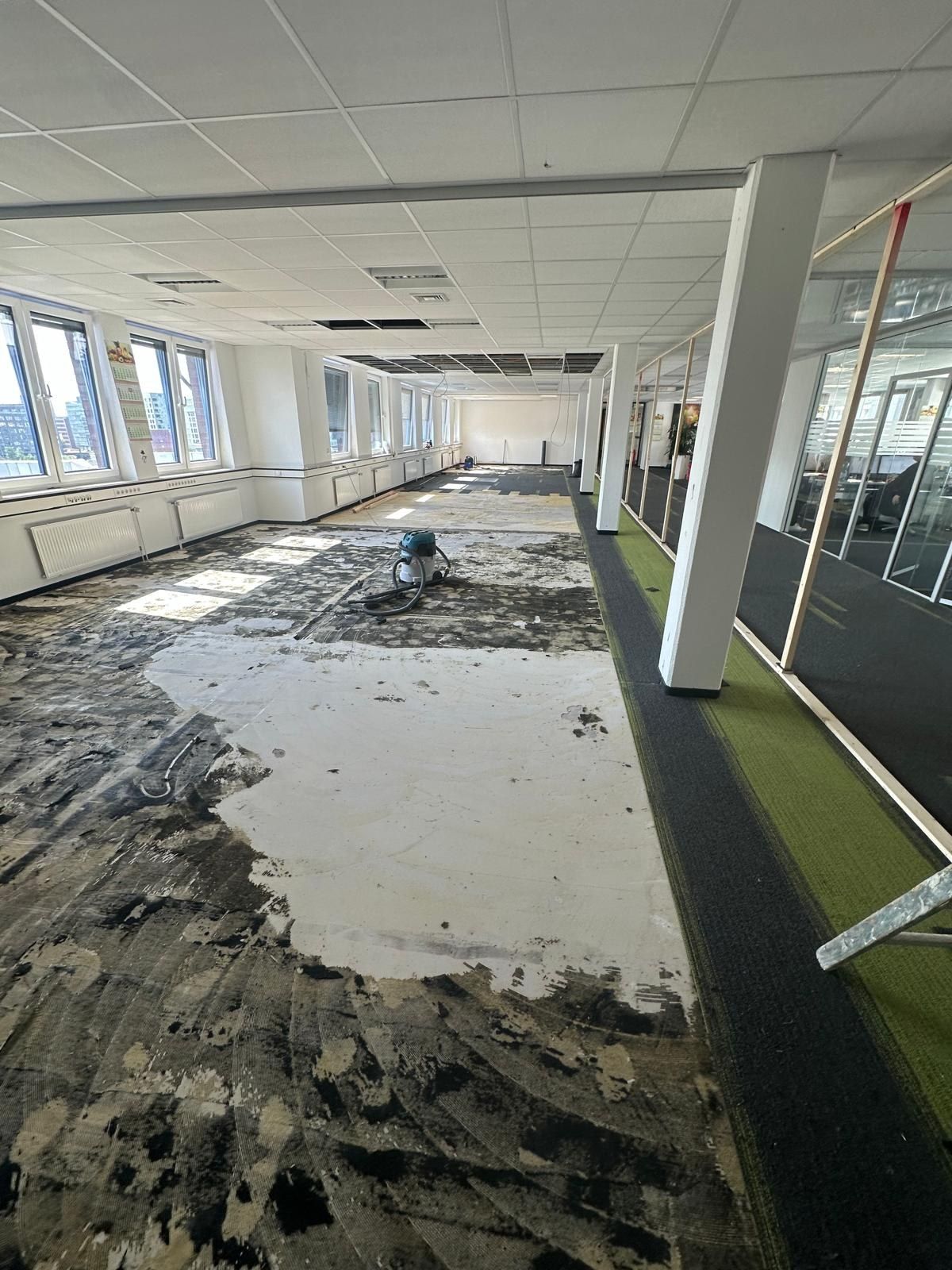 Bürofläche im Zwischenstand: Altbelag abgezogen, Kleberreste auf dem Estrich, ein Schleif- und Absauggerät steht in der Fläche, Fensterband und Rasterdecke bleiben unangetastet.