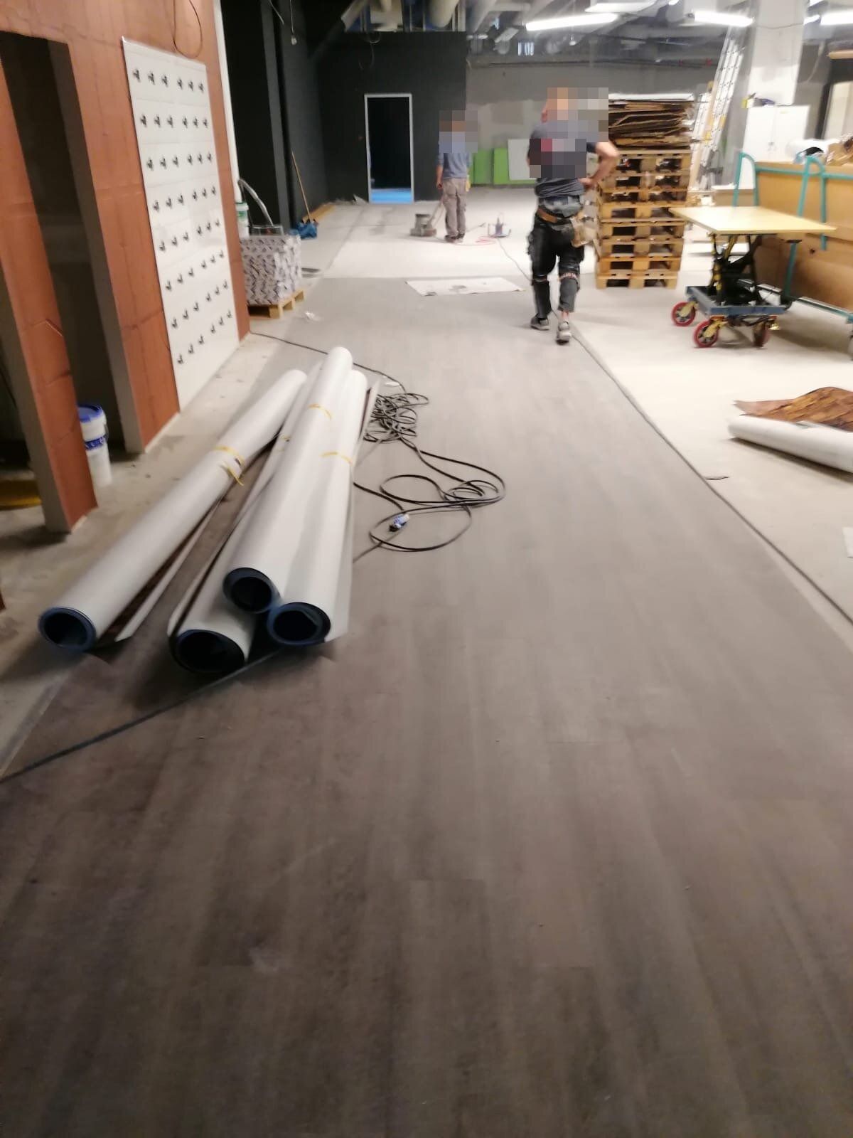 Große Halle im Ausbau: auf der einen Hälfte liegt dunkler Plankenbelag, daneben offener Estrich; im Vordergrund aufgerollte Bahnenware und ein Kabelring, im Hintergrund Paletten und ein Hubtischwagen; zwei Handwerker von hinten, Gesichter und Aufdruck unkenntlich gemacht.