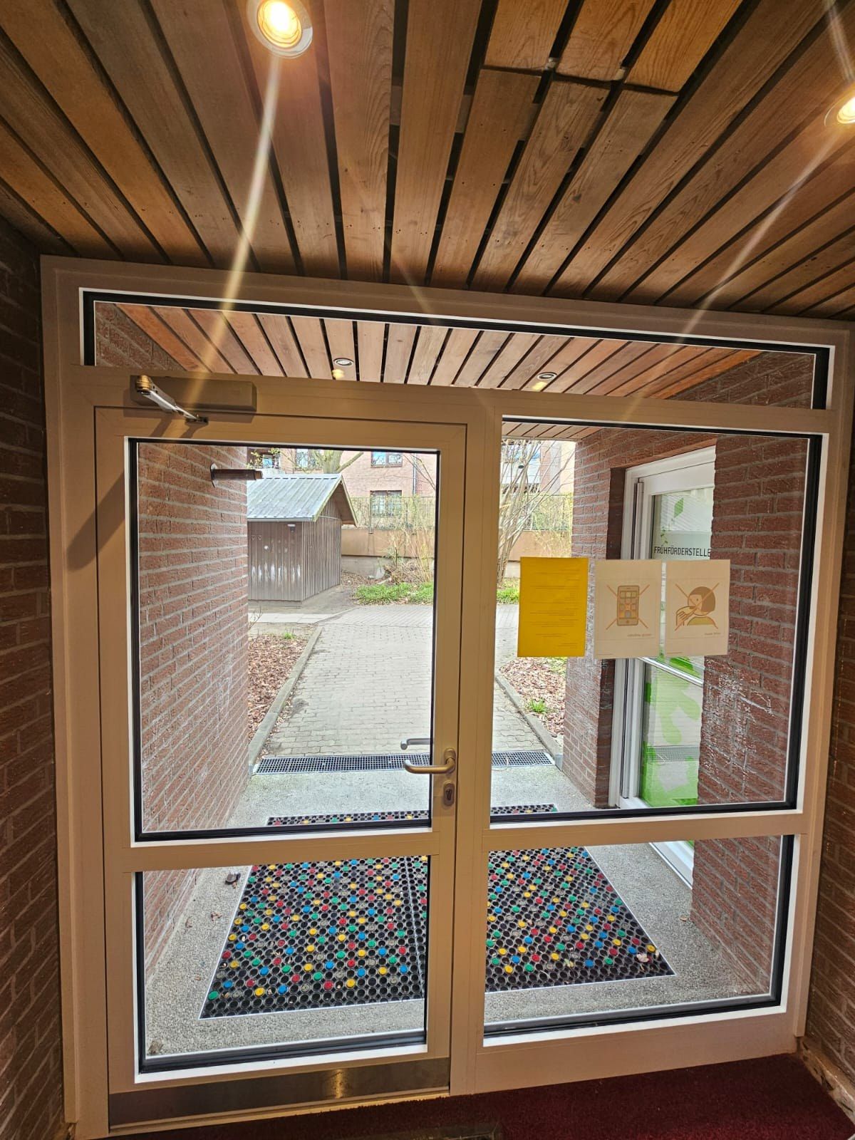 Neue Eingangstür einer Kita von innen: zweiflügelige Aluminiumkonstruktion mit großen Glasfeldern und Oberlicht, obenliegender Türschließer, dahinter der gepflasterte Vorplatz.