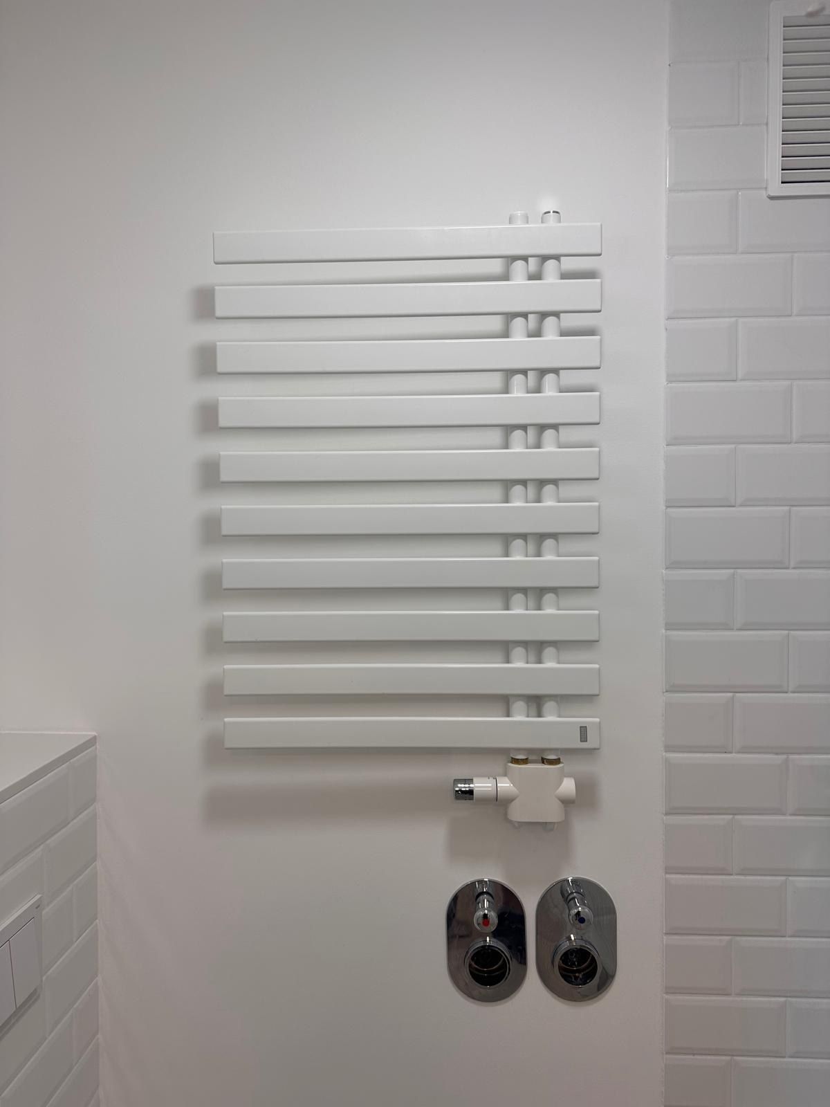 Weißer Handtuchheizkörper an einer glatten Wand neben weißen Metrofliesen, darunter zwei verchromte Anschlussrosetten und das Thermostatventil.