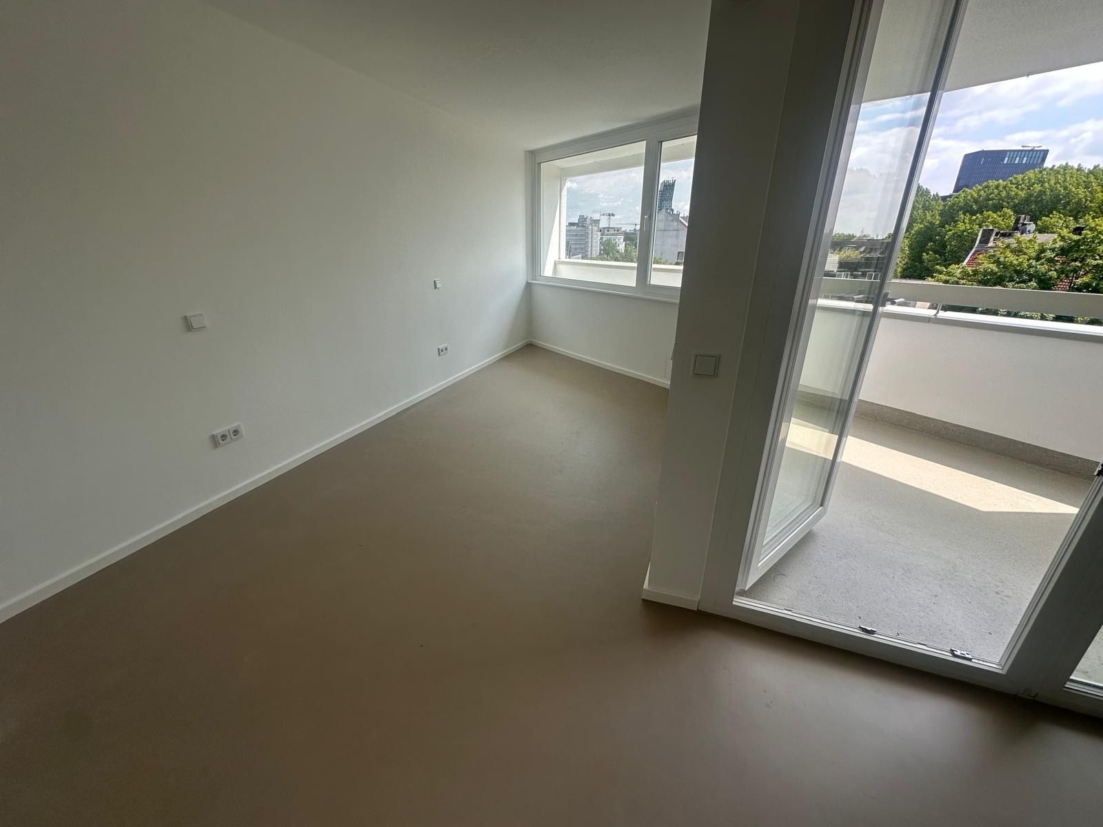 Fertiges Zimmer mit neuem Eckfenster und geöffneter Balkontür, heller Bodenbelag, weiße Wände, Blick ins Grüne und auf ein Hochhaus.