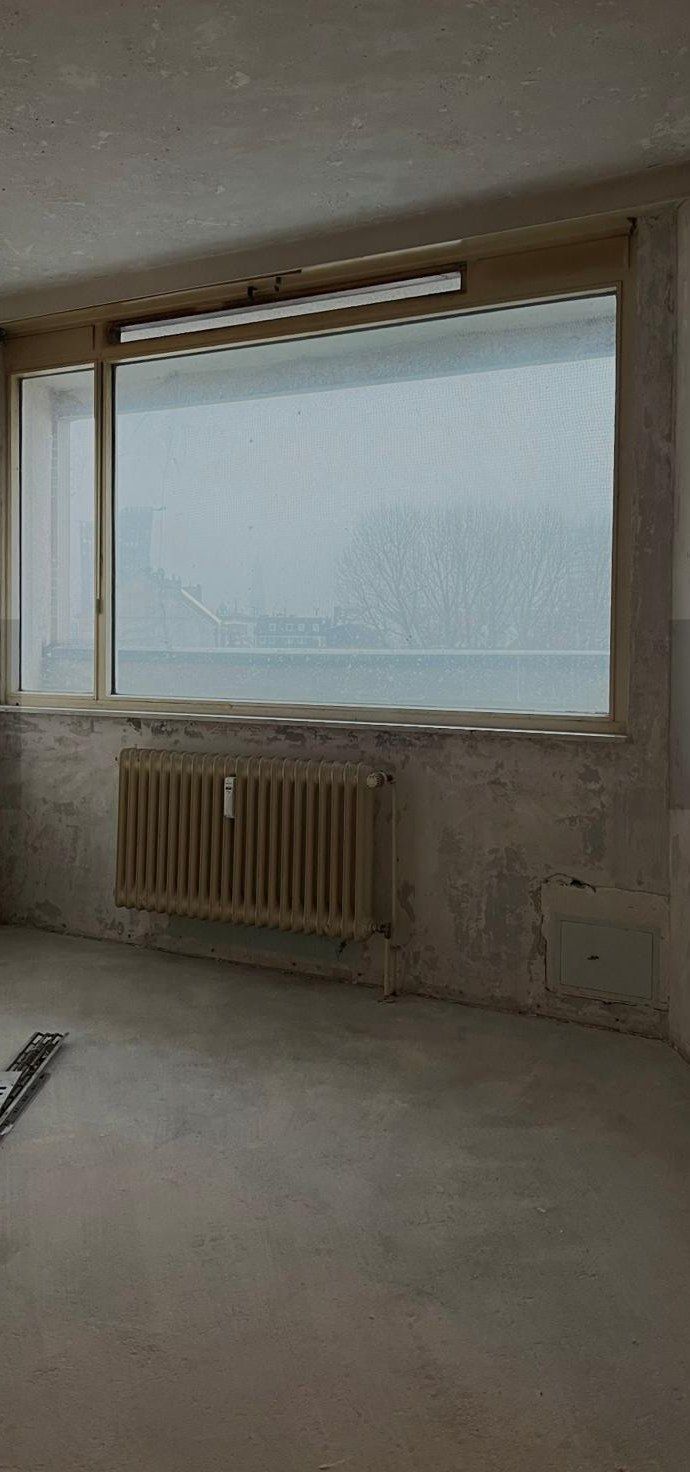 Zimmer vor der Sanierung: abgeschlagener Putz an der Brüstung, altes Fensterband mit Gussheizkörper darunter, nackter Estrich, Blick auf den Hafen.