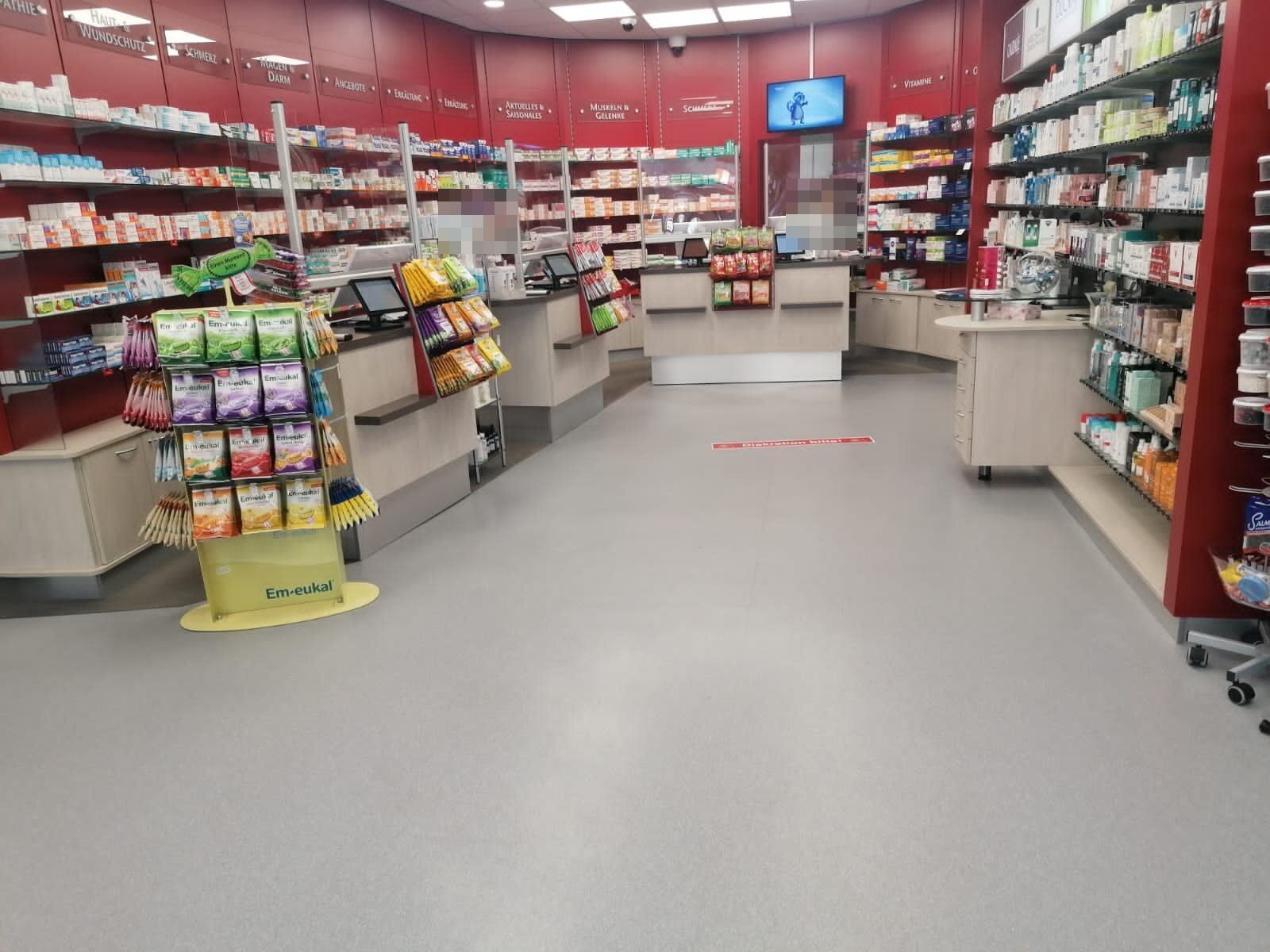 Verkaufsraum einer Apotheke mit rotem Wandregal, freistehender Beratungstheke und hellgrauem Bodenbelag; im Hintergrund zwei unkenntlich gemachte Personen.