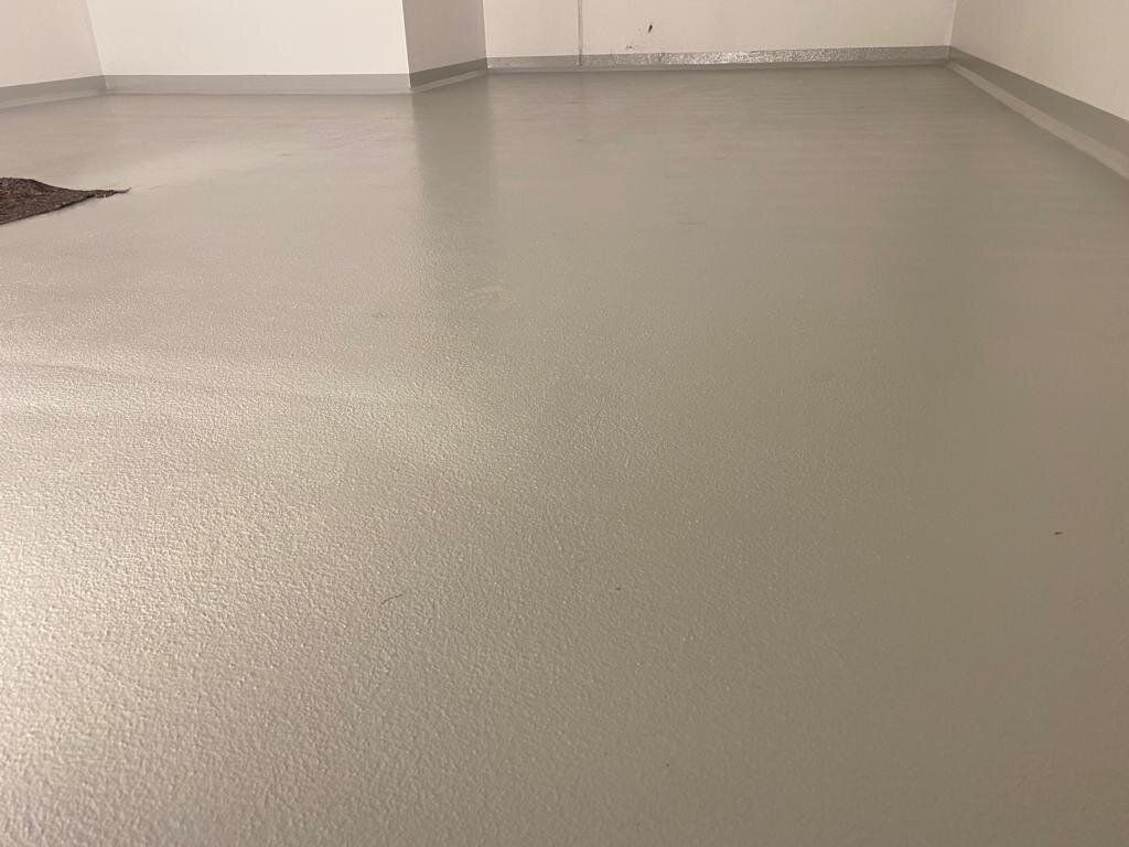 Leerer Verkaufsraum mit fugenloser grauer Bodenbeschichtung, grau hochgezogenem Wandsockel und frisch gestrichenen weißen Wänden; am linken Bildrand eine offene Stelle im Untergrund.
