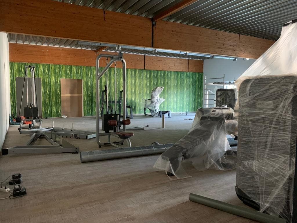 Trainingsfläche im Umbau: teilweise verlegter Boden in Holzoptik, Trainingsgeräte unter Schutzfolie, grüne Fototapete an der Rückwand, Decke aus Holzbindern und Trapezblech, im Hintergrund ein Gerüstturm.