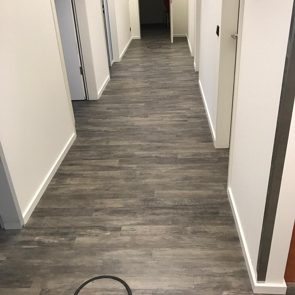 Flur einer Büroetage mit grauem Designboden in Holzoptik, weißen Türen und Sockelleisten, einer runden Bodendose im Belag und einem aufgerollten Kabelstrang am Boden.