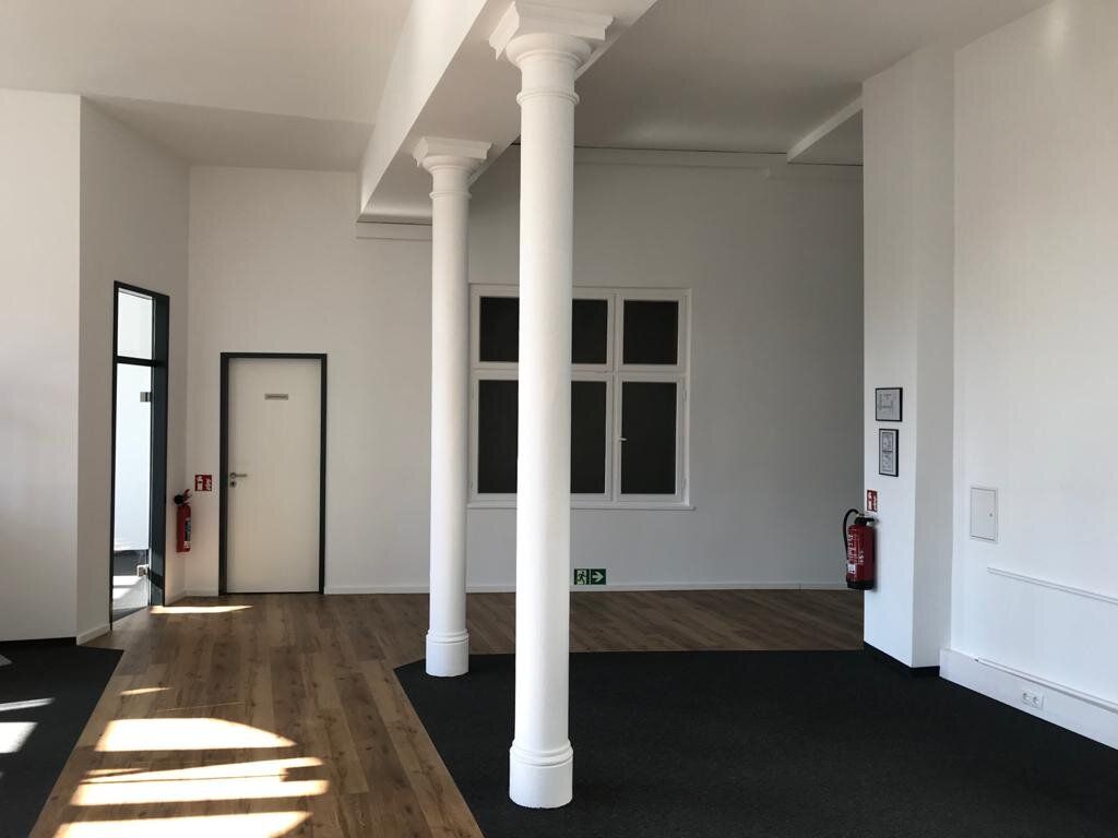 Leerer Büroraum mit zwei schlanken weißen Säulen mit Kapitell, Sprossenfenster, Dielenboden in Holzoptik neben dunklem Teppichbelag, Feuerlöscher und Rettungszeichen an der Wand.
