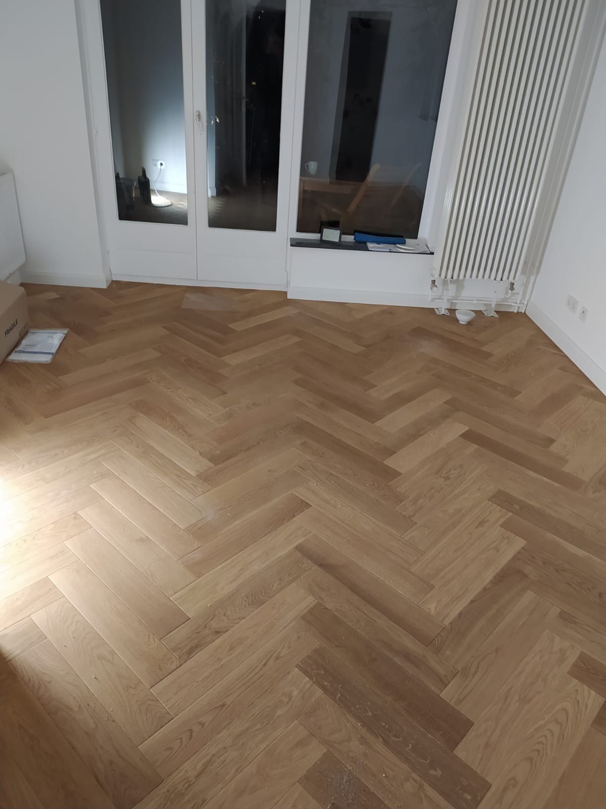 Derselbe Raum fertig: helles Fischgrätparkett bis an die weiße Fenstertür, Röhrenheizkörper an der Wand, Fensterbank und Zargen weiß gestrichen.