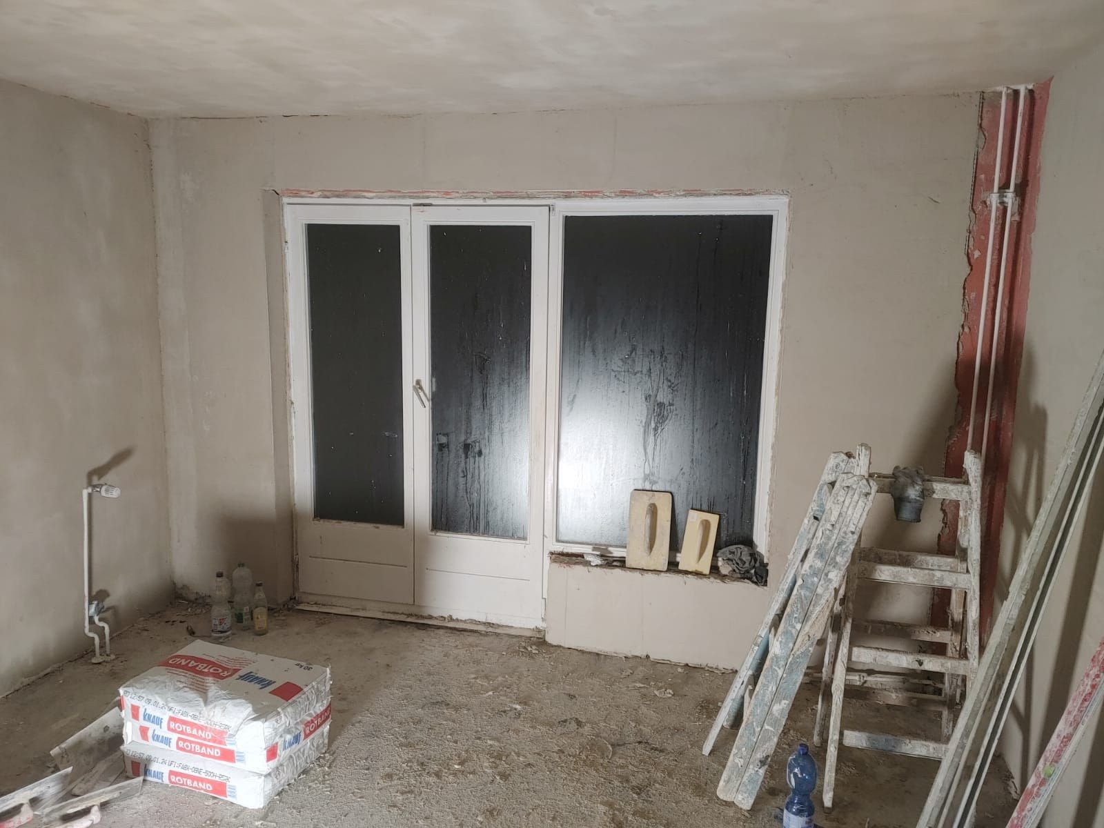 Wohnraum im Rohzustand: verputzte Wände ohne Anstrich, alte weiße Fenstertür mit dunklen Scheiben, Leiter, Putzsäcke und Werkzeug auf dem nackten Boden.