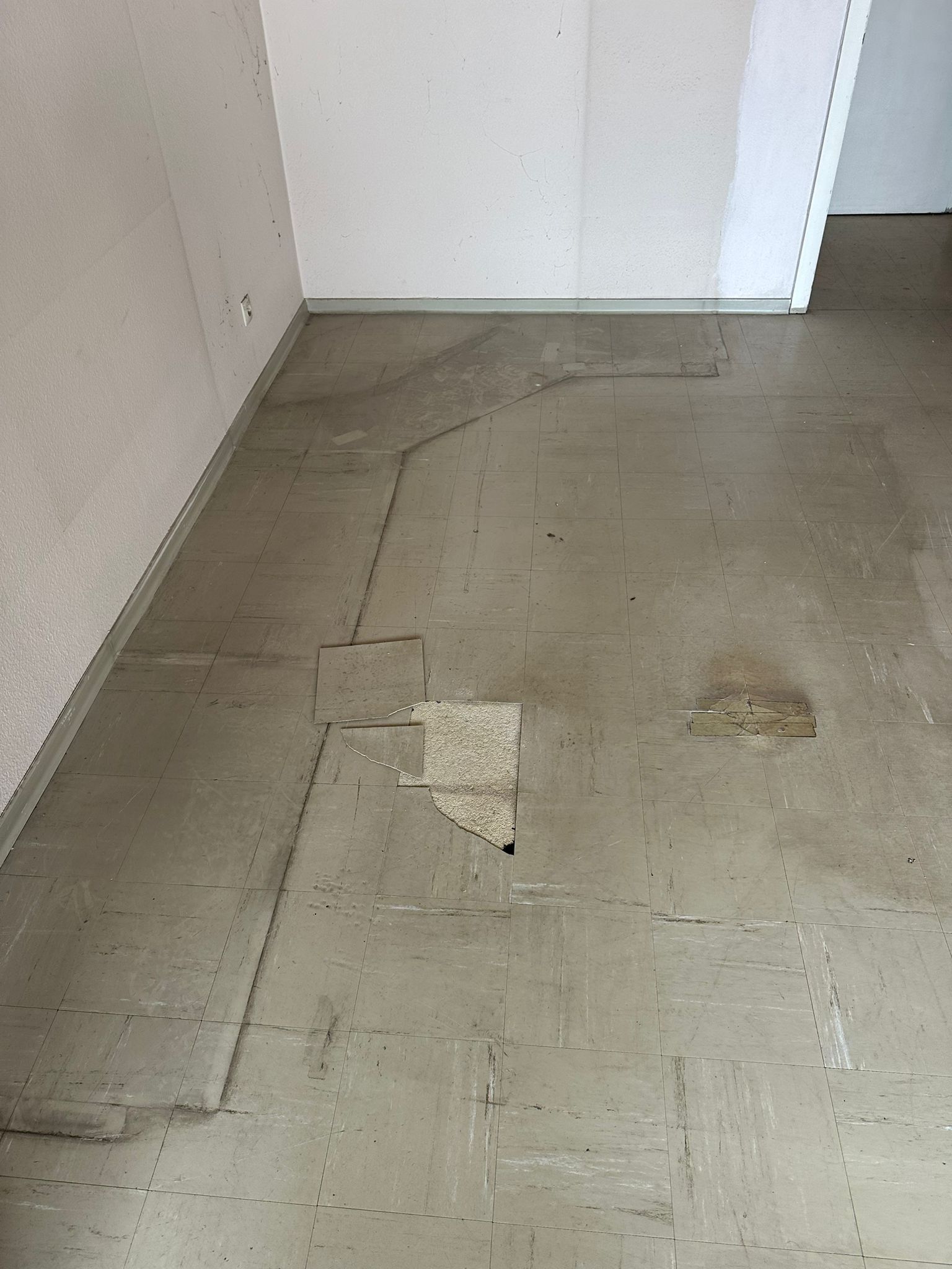 Alter Bodenbelag vor dem Rückbau: verklebte helle Fliesen mit ausgebrochenen Stellen und Klebstoffresten, aufgebogene Kante an der Wand.