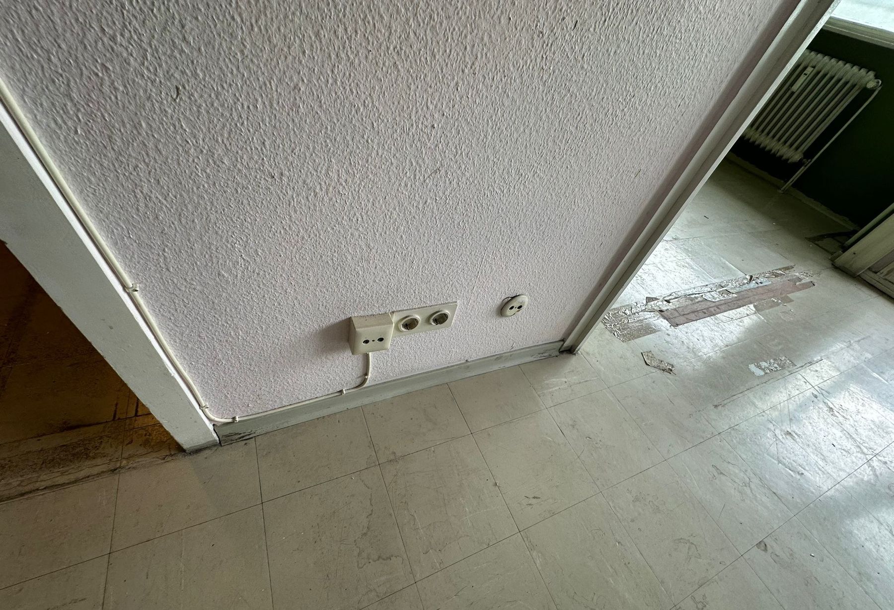 Elektroinstallation vor der Sanierung: Steckdosen und Leitungen auf Putz an einer Raufasertapete, daneben der alte Fliesenboden mit Klebstoffresten.