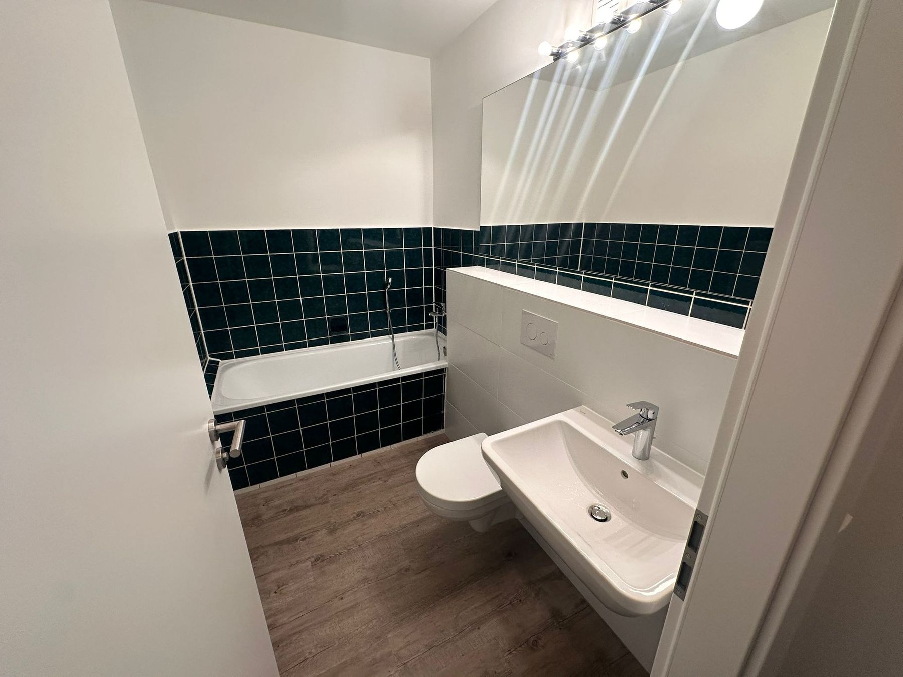 Neues Bad: weiße Wände mit einem umlaufenden Band aus dunkelgrünen Fliesen, Wanne mit gefliester Schürze, Wand-WC, Waschtisch mit durchlaufender Ablage und breitem Spiegel.