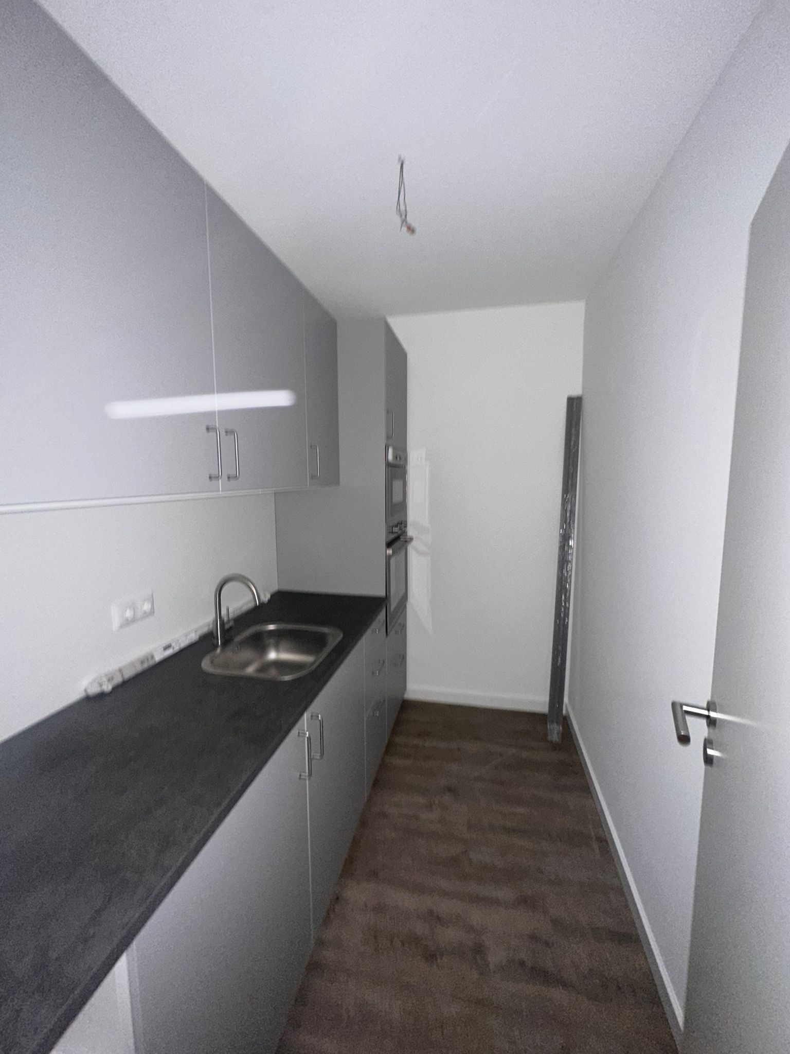 Neue Küchenzeile in Grau mit dunkler Arbeitsplatte, Edelstahlspüle, Einbaubackofen und Hängeschränken, davor durchlaufender Vinylboden.