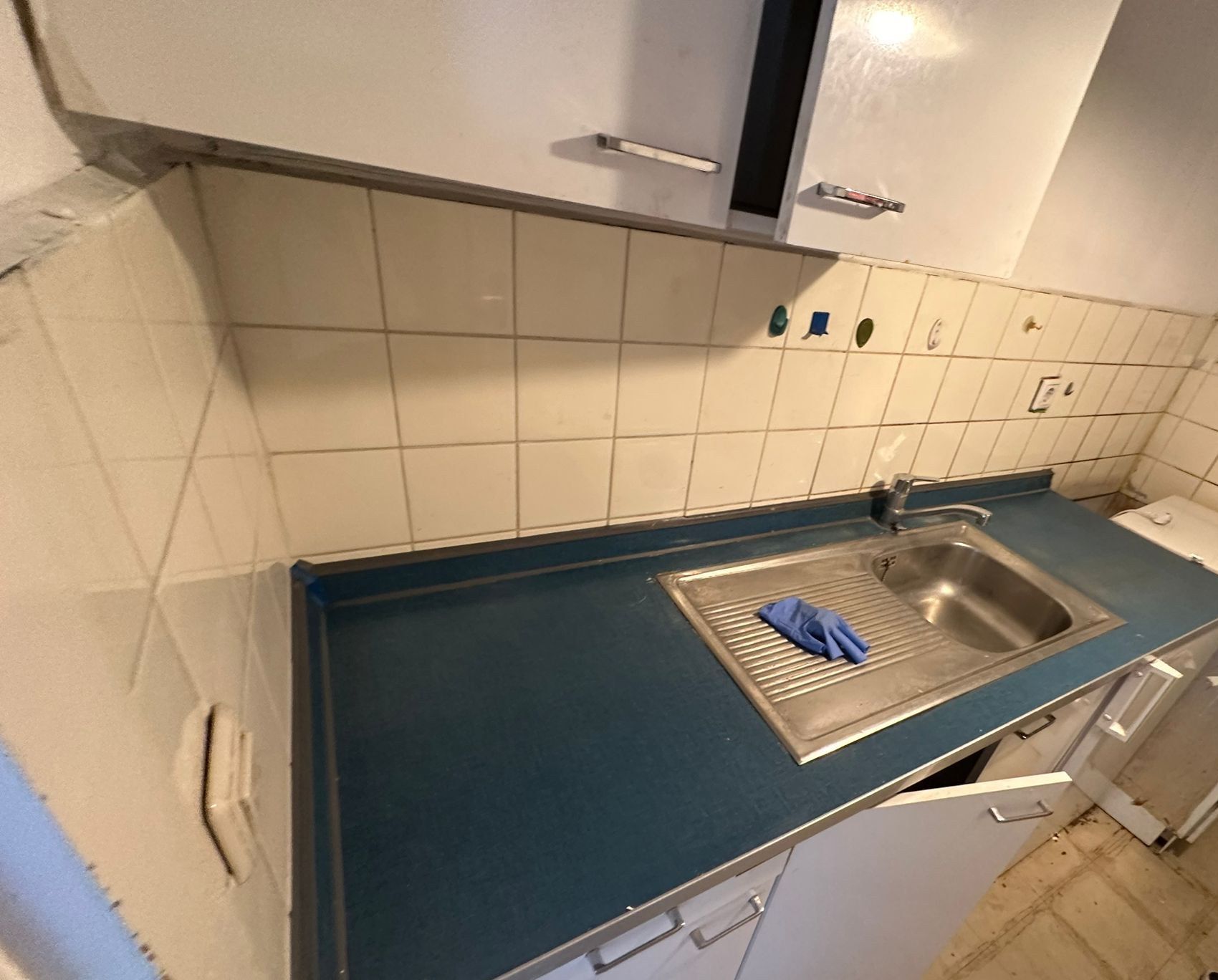 Alte Küchenzeile: blaue Kunststoffarbeitsplatte mit Edelstahlspüle, weiße Fliesen als Spritzschutz, Hängeschränke, ausgebauter Unterschrank.