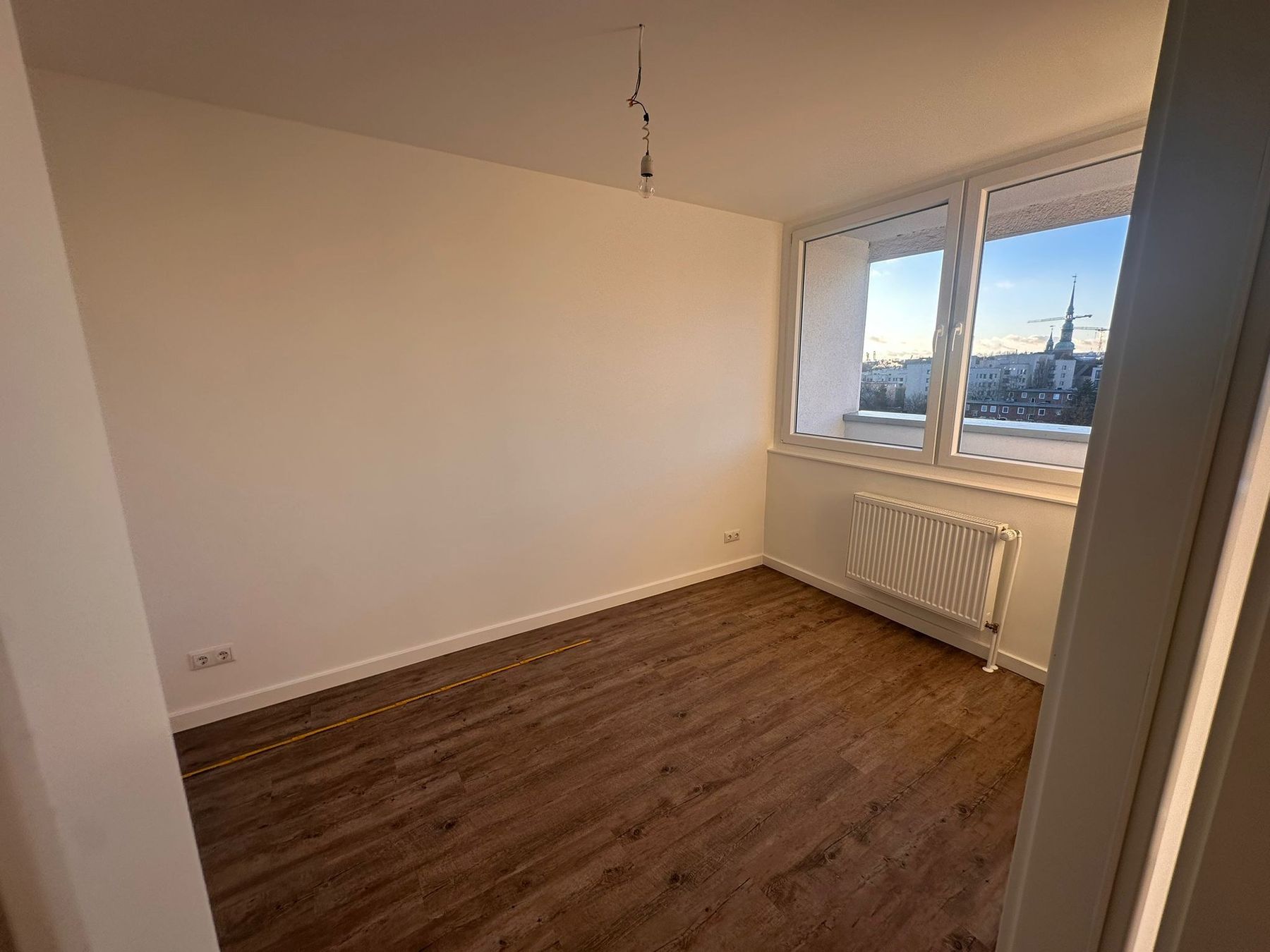 Fertiges Schlafzimmer mit dunklem Vinylboden, weißen Wänden, Flachheizkörper und Fenster mit Blick auf die Nachbarbebauung.