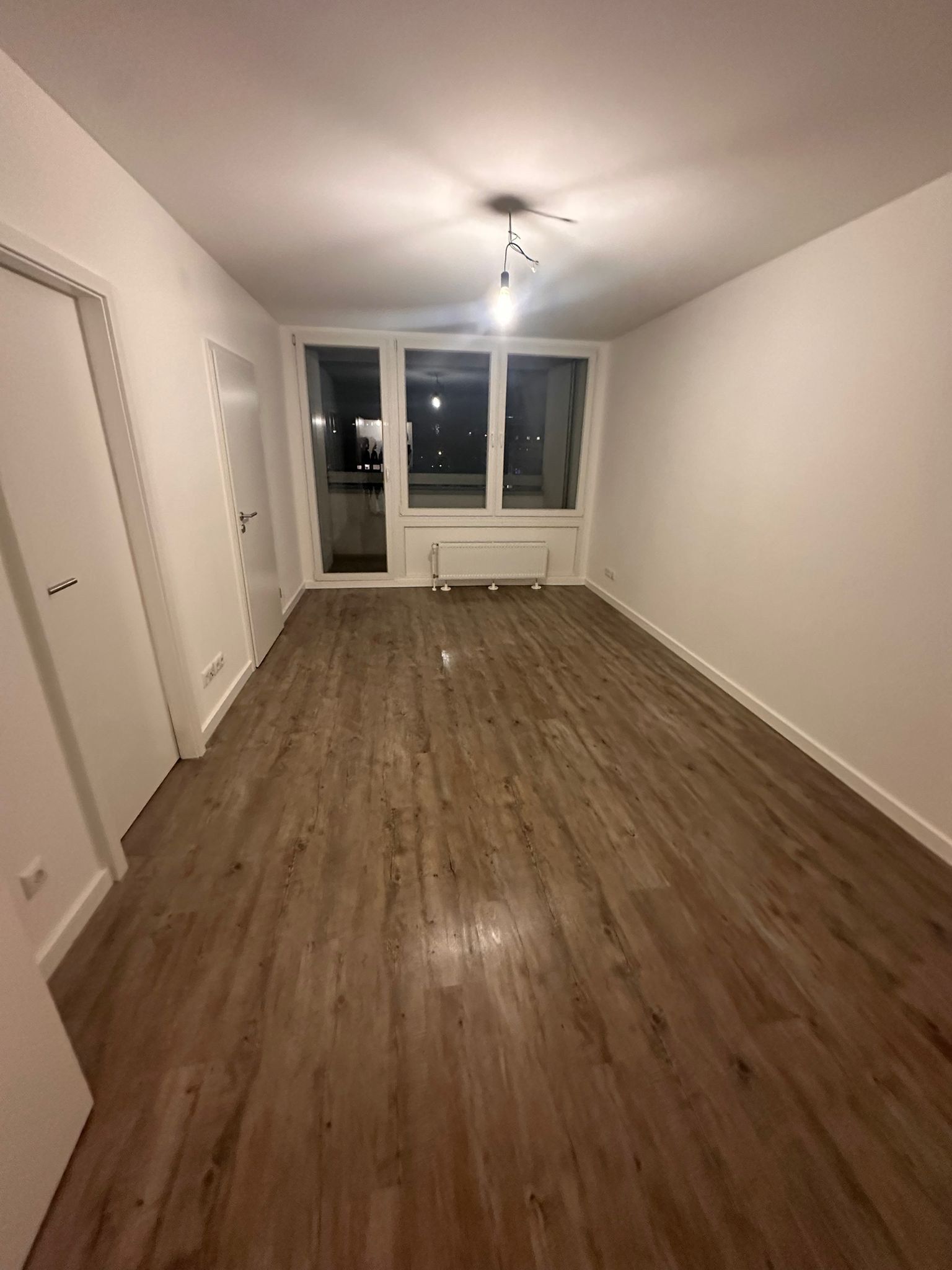 Fertiger Raum mit durchgehendem Vinylboden in dunkler Holzoptik, Sockelleisten gesetzt, Deckenleuchte an provisorischer Fassung.
