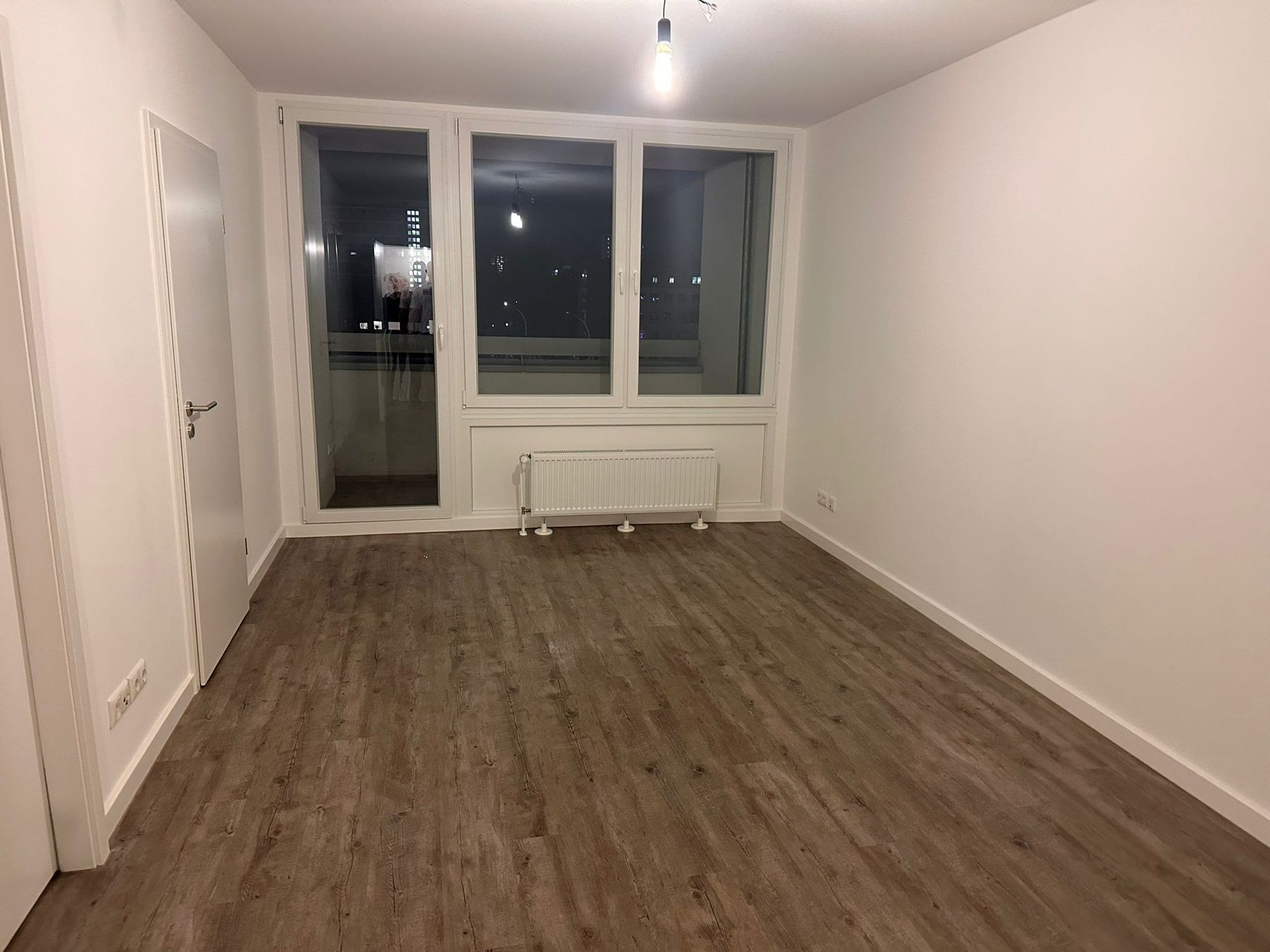 Fertiger Wohnraum bei Nacht: dunkler Vinylboden, weiße Wände, Flachheizkörper unter der Fensterfront, Tür zum Flur geöffnet.
