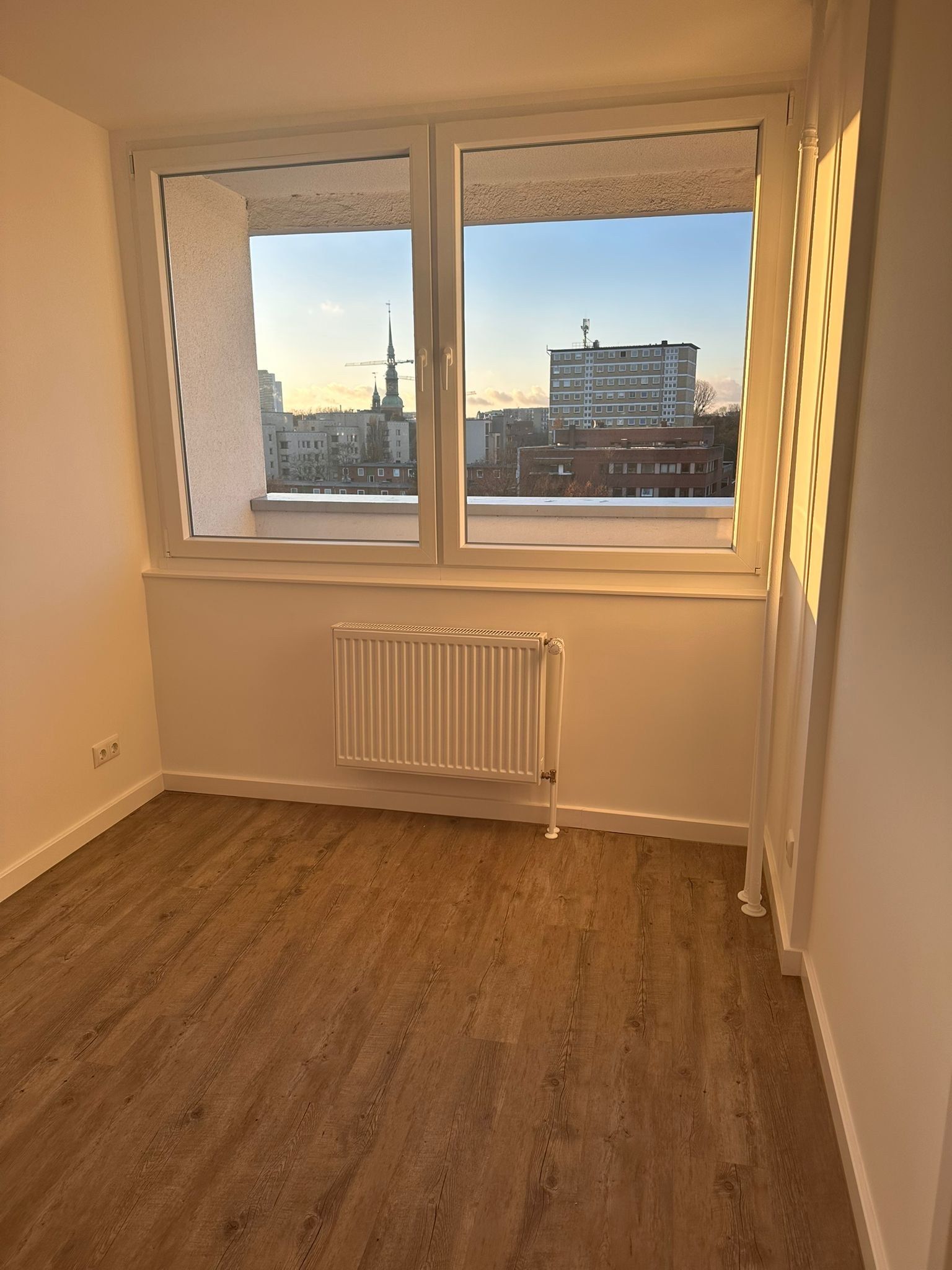 Fertiges Zimmer im Abendlicht: dunkler Vinylboden in Holzoptik, weiße Wände, neues Fenster mit Heizkörper darunter, Blick über die Dächer zum Turm der Michaeliskirche.