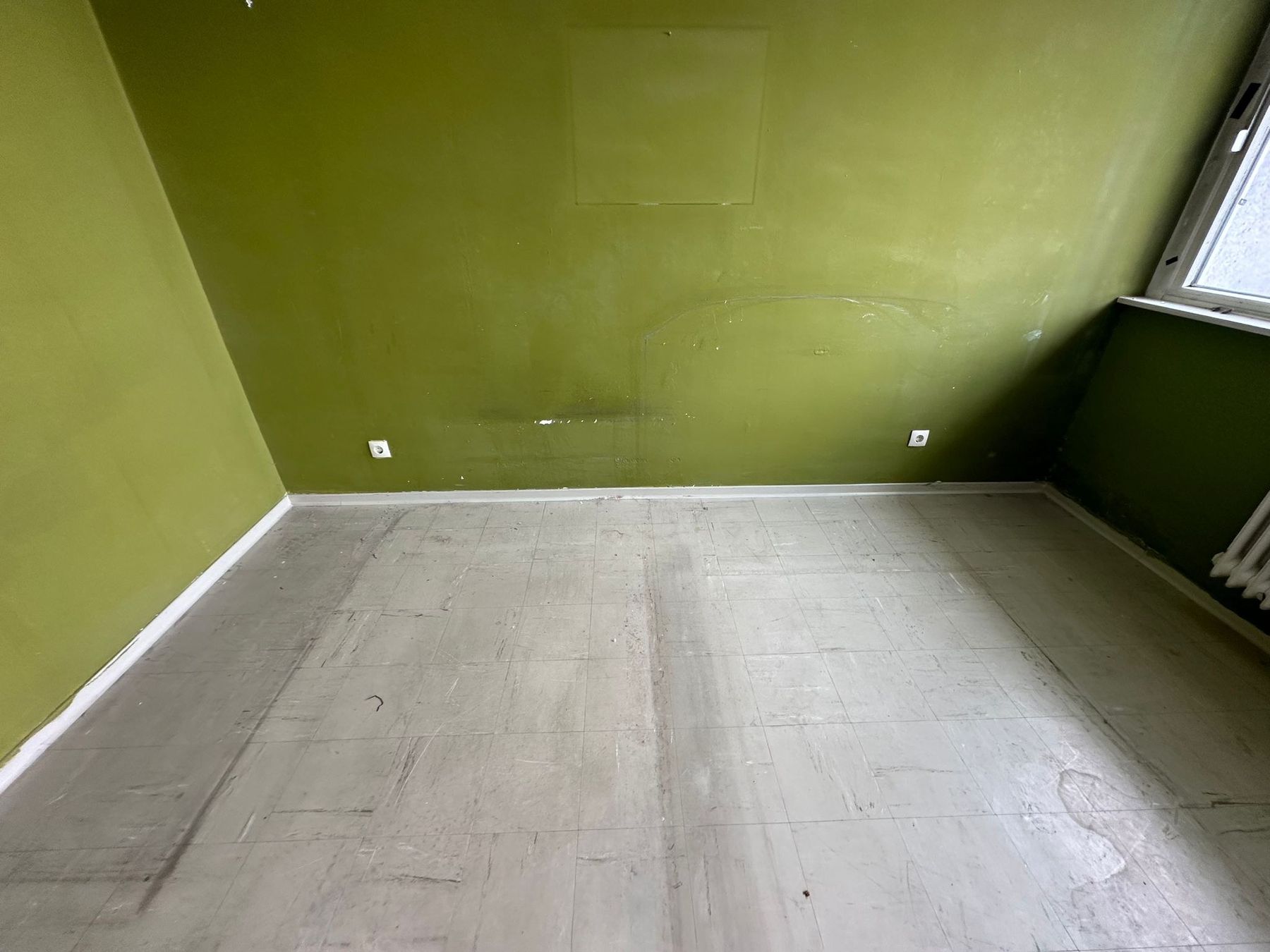 Zimmer vor der Sanierung: olivgrün gestrichene Wände mit ausgebesserten Stellen, alter grauer Fliesenbelag am Boden, Heizkörper unter dem Fenster.