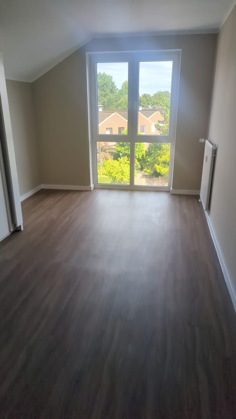 Fertiges Zimmer unter der Dachschräge: Vinylboden in dunkler Holzoptik, warmgraue Wände, bodentiefes Fenster mit Blick auf Reihenhäuser und Gärten.