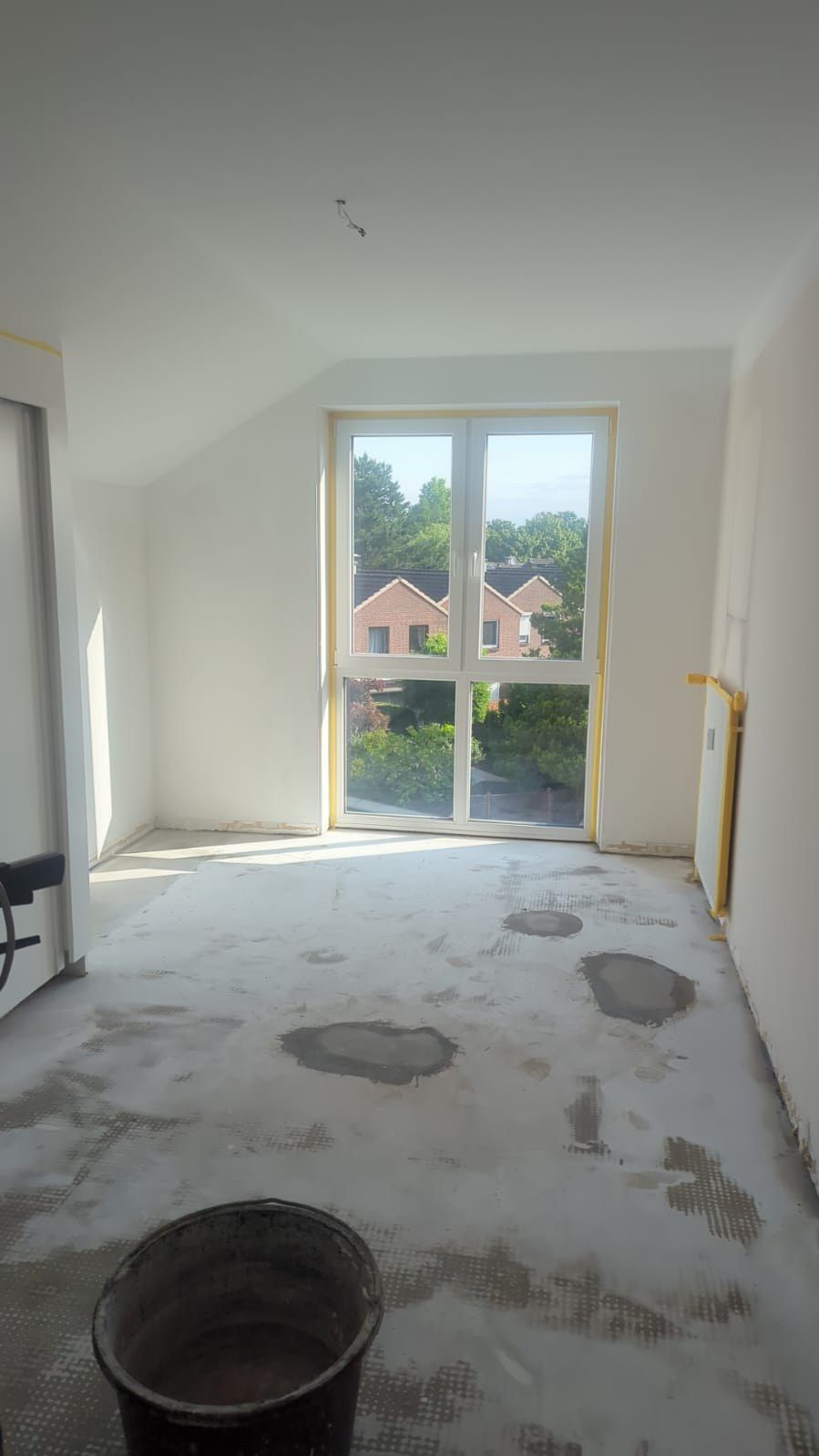 Zimmer unter der Dachschräge im Rohbau: offener Estrich mit Spachtelresten, ungestrichene Wände, abgeklebter Fensterrahmen, Eimer in der Raummitte.