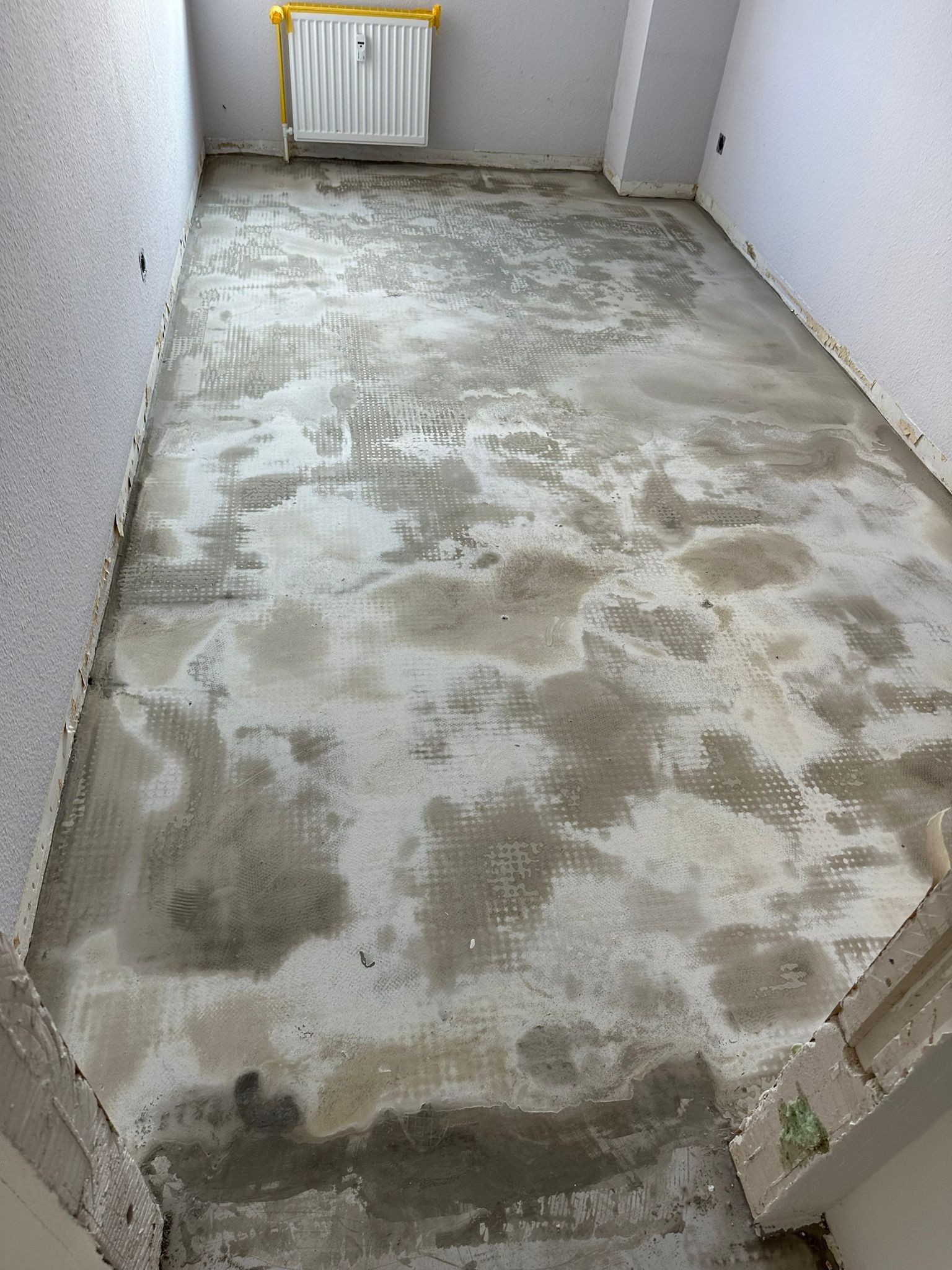 Estrich vor dem Belag: geschliffene Fläche mit Resten von Kleber und Spachtelmasse, Heizkörper an der Wand, Wandanschlüsse offen.