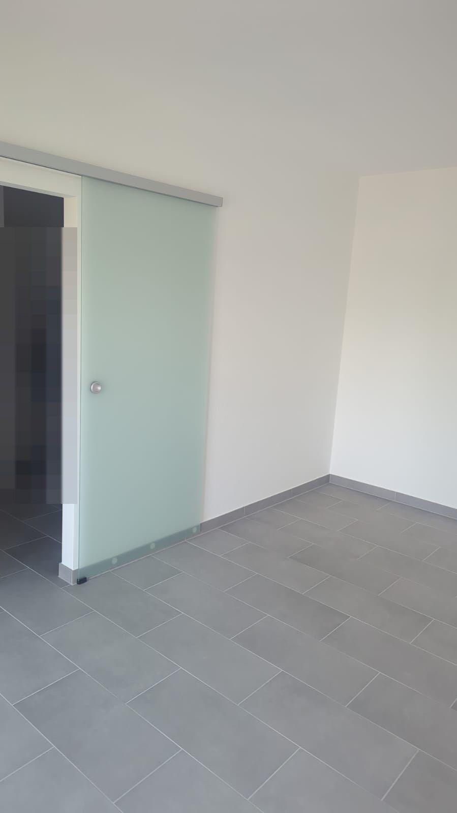 Glasschiebetür aus mattem Sicherheitsglas vor einer Wandöffnung, geführt an einer Laufschiene, davor grauer Fliesenboden.