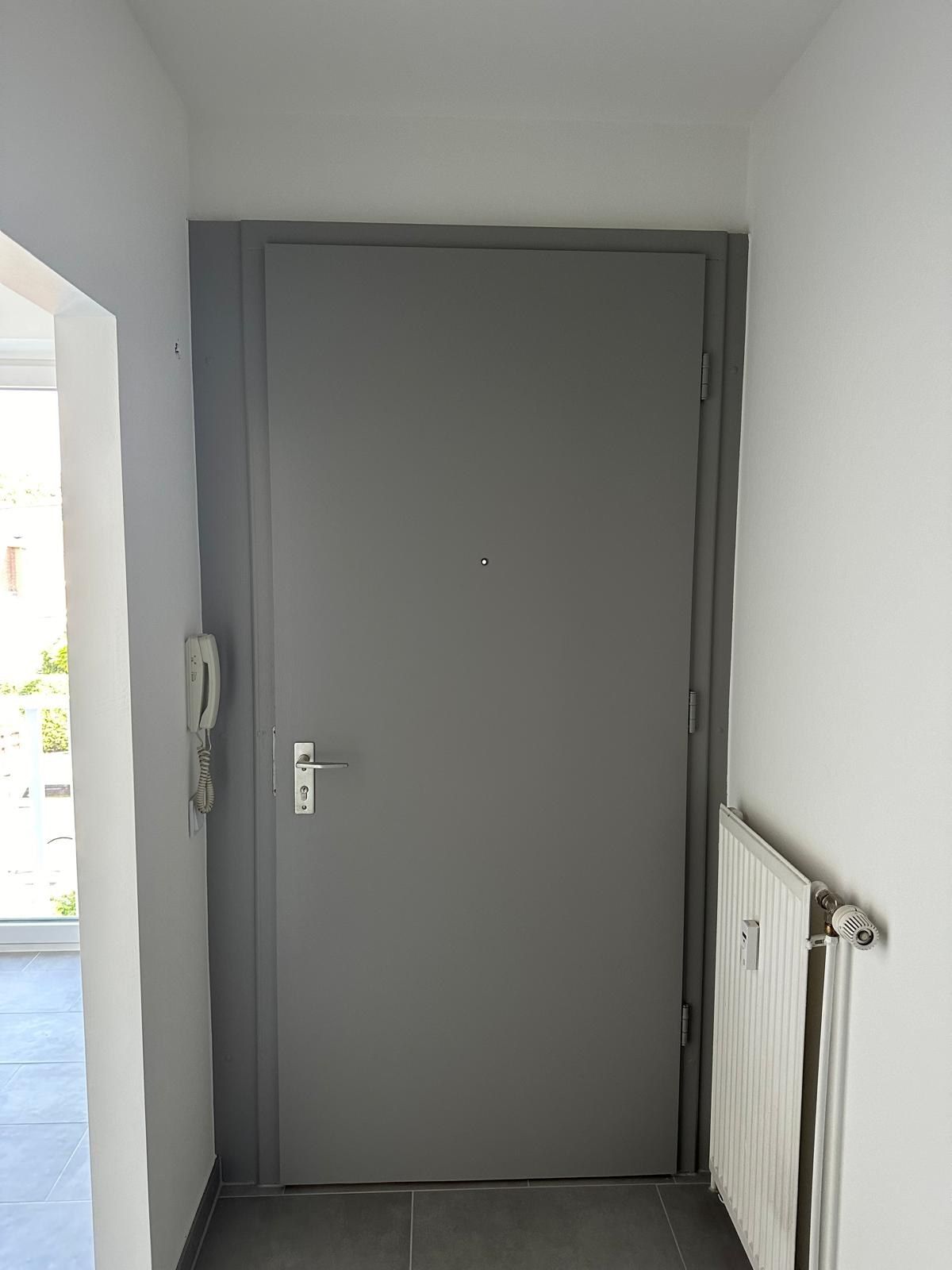 Neue Wohnungseingangstür in Grau mit umlaufender Zarge, Türspion und Edelstahlklinke, links an der Wand die Gegensprechstelle, rechts ein Heizkörper.