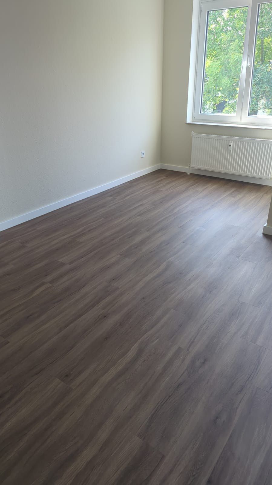 Dasselbe Zimmer fertig: Vinylboden in Holzoptik, hell gestrichene Wände mit weißer Sockelleiste, Flachheizkörper unter dem Fenster.