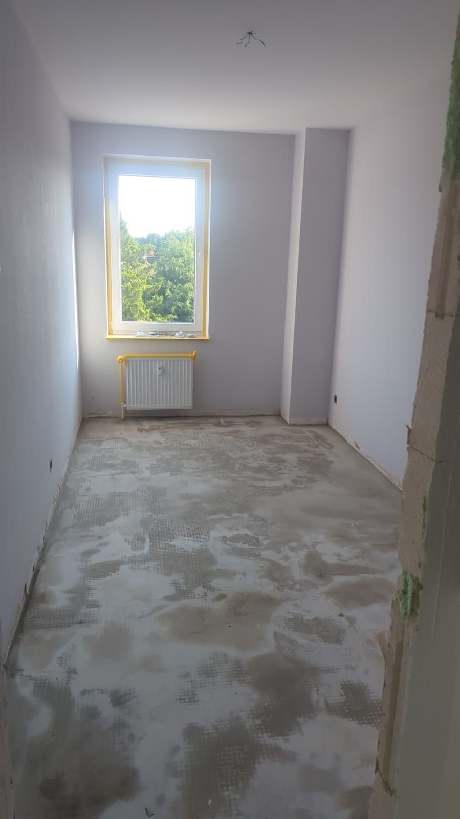 Zimmer im Rohbau: gespachtelter Estrich, verputzte Wände ohne Anstrich, Heizkörper unter dem abgeklebten Fenster, offene Wandkante zum Flur.